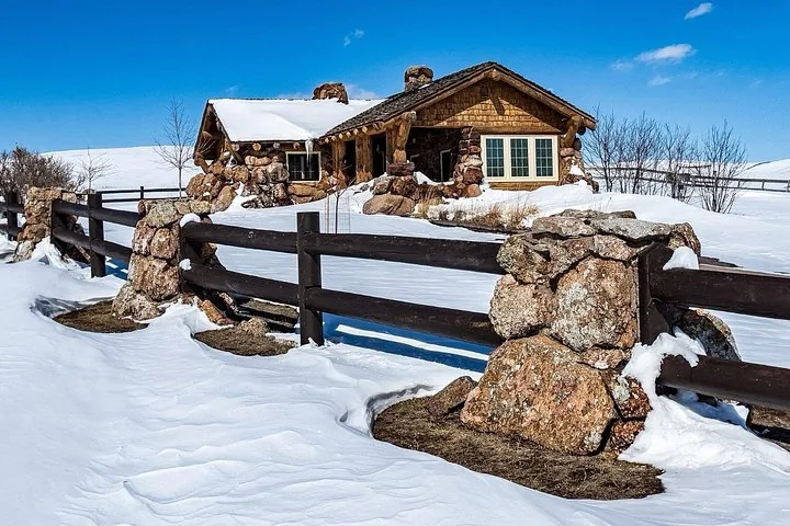 Blockhütte aus Holz und Stein hinter einem Holzzaun in verschneiter Landschaft